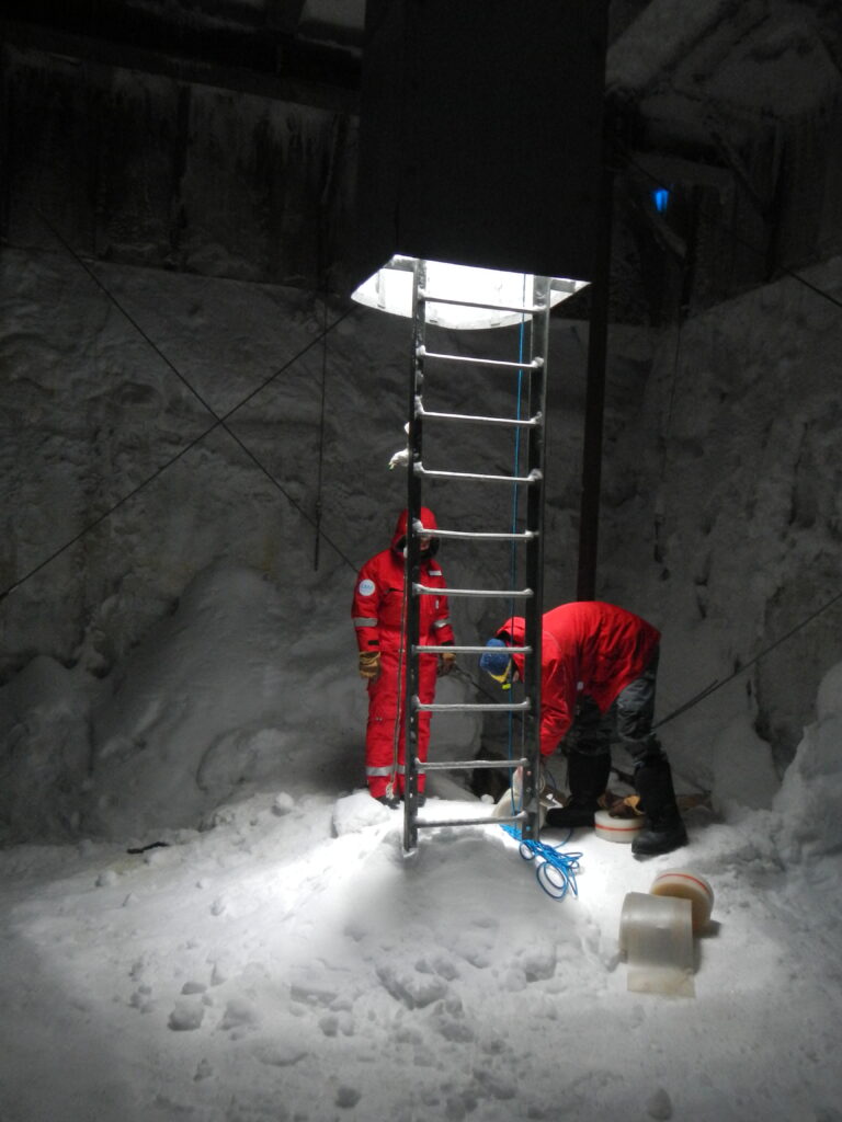 Snowy cave. Two humans in read standing by a ladder leading up to the light.