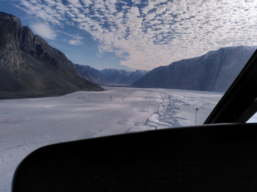 View from helicopter cockpit of glacier surrounded by tall mountains.